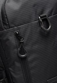 Sac à dos noir en tissu texturé avec un motif en grille. Comprend une fermeture éclair noire et un tirette, ainsi qu'un clip de fixation en métal.