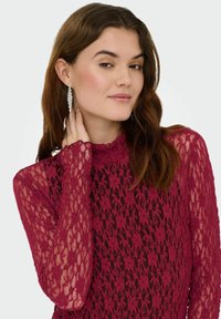 Haut en dentelle bordeaux avec manches longues et col montant. Motif floral texturé, accentué par des boucles d'oreilles pendantes étincelantes sur fond clair.