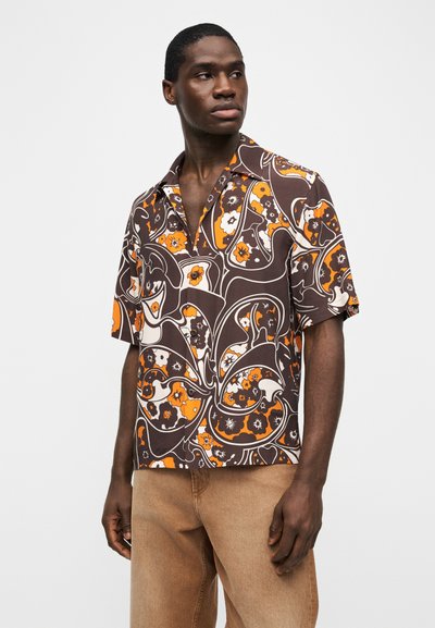 Νέος άνδρας που φορά καφέ, πορτοκαλί και λευκό πουκάμισο με κοντά μανίκια και floral μοτίβο και μπεζ παντελόνι, κοιτάζει προς τα πλάγια πάνω σε απλό φόντο.