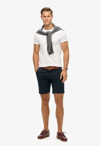 Hvid kortærmet t-shirt, navy shorts, grå sweater draperet over skuldrene, brun bælte og burgunder bådsko. Enkel og afslappet påklædning.