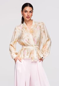 Femme portant un chemisier portefeuille en soie à motifs floraux avec des manches bouffantes et une ceinture à la taille, associé à un pantalon large taille haute de couleur rose pâle.