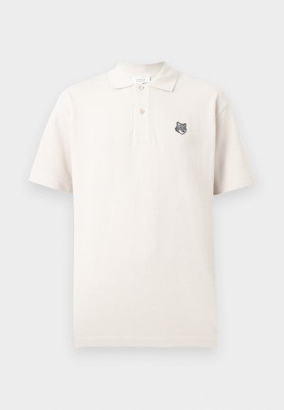 Κοντομάνικο κρεμ πόλο πουκάμισο με δύο κουμπιά και ένα μικρό κεντημένο κεφάλι λύκου στην αριστερή πλευρά του στήθους.