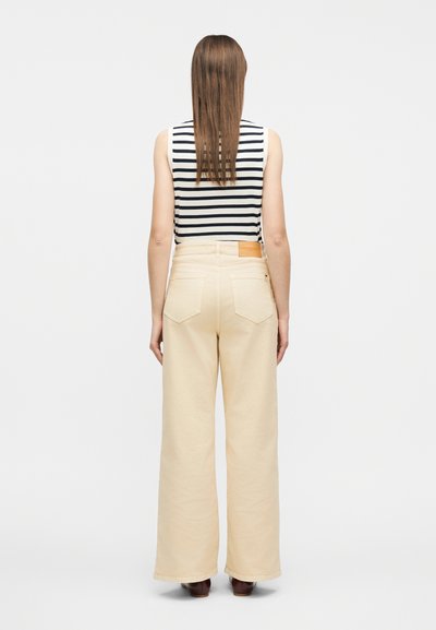 Γυναίκα που στέκεται με την πλάτη προς τα εμάς, φορώντας αμάνικη μαύρη και λευκή ριγέ μπλούζα, μπεζ παντελόνι με φαρδιά πόδια και ψηλή μέση, και καφέ παπούτσια.