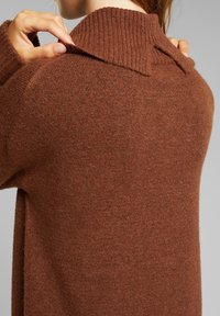 Maglione lavorato a maglia marrone con colletto alto a coste e dettagli sottili nella parte posteriore del colletto, realizzato in un morbido materiale testurizzato.