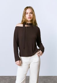 Haut marron à manches longues avec un design décolleté et un col noué. Associé à un pantalon blanc taille haute et à une texture lisse.