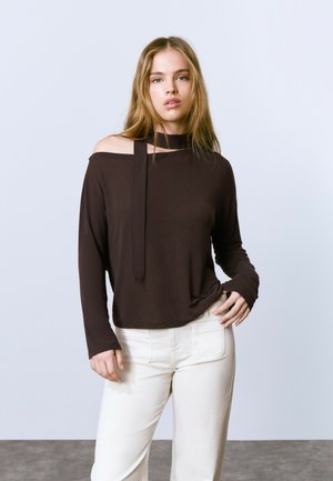 Top marrón de manga larga con un diseño off-the-shoulder y un collar atado. Combinado con pantalones blancos de cintura alta y una textura suave.