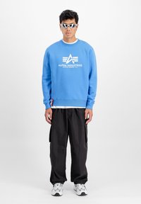 Plava dukserica s bijelim logotipom "Alpha Industries", uparena s crnim cargo hlačama i bijelim tenisicama. Model nosi bijele sunčane naočale.