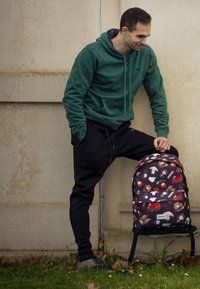 Mochila negra y con estampado de personajes de dibujos animados; correas de tela, doble cremallera y un bolsillo delantero. Sudadera verde y pantalones de chándal negros.