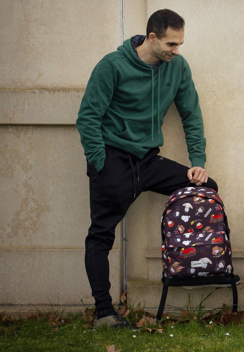 Mochila negra y con estampado de personajes de dibujos animados; correas de tela, doble cremallera y un bolsillo delantero. Sudadera verde y pantalones de chándal negros.