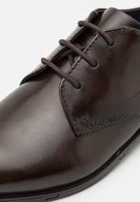 Chaussure de ville en cuir marron avec une texture lisse, un bout arrondi et des accents cousus. Présente un design à lacets et une brillance subtile.