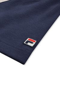 Navyblåt stofhjørne med en lille firkantet lap med et rødt og sort stiliseret bogstav på en hvid baggrund.