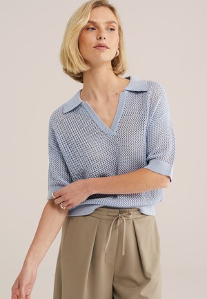 Szőke nő világoskék hálós kötött galléros rövid ujjú felsőt és bézs megkötős nadrágot viselve, karját keresztbe téve pózol.