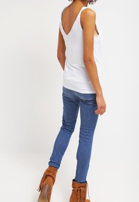 Top bianco senza maniche con una profonda scollatura a V sul retro, abbinato a jeans blu aderenti e stivaletti alla caviglia marroni con frange. Design e texture semplici e casual.