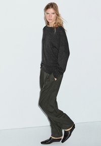 Dunkelgrauer, langärmliger Pullover, entspannte Passform, kombiniert mit olivgrünen, weit geschnittenen Hosen. Schwarze, spitze Ballerinas runden das Ensemble ab.