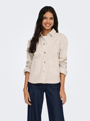 Femme souriante aux longs cheveux foncés portant une veste beige clair boutonnée avec des poches et un jean large bleu foncé sur un fond uni.