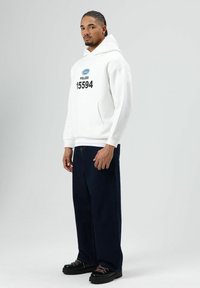 Witte hoodie met "POLIZEI 15594" in het zwart gedrukt, gecombineerd met donkerblauwe wijde jeans en zwarte veterboots. Gladde stoftextuur.