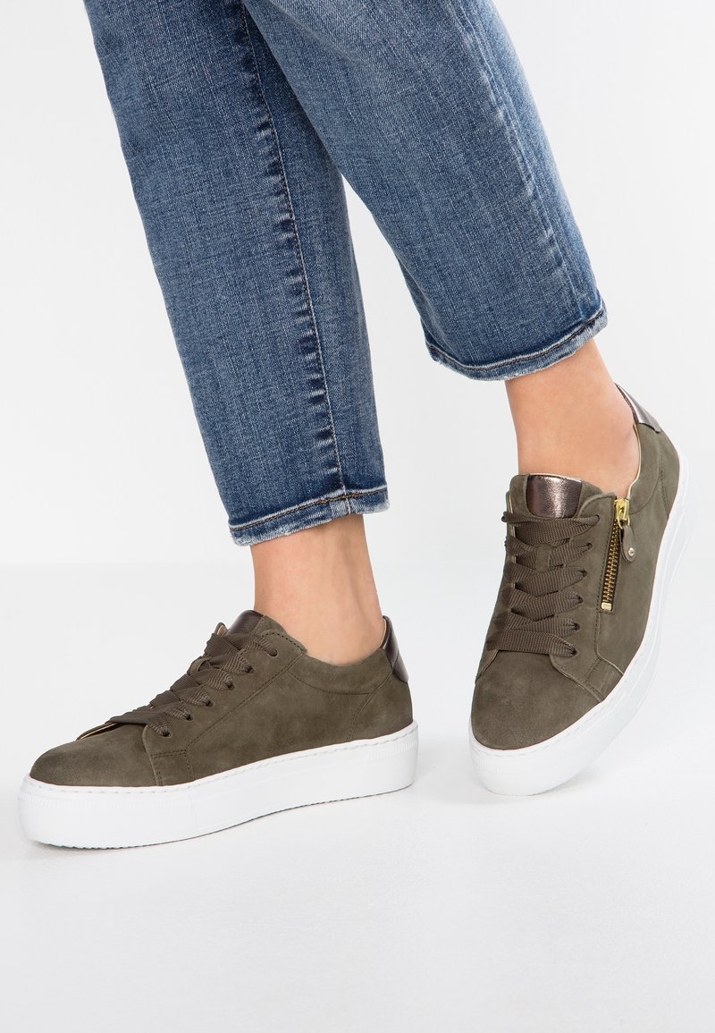 Oliwkowe zamszowe sneakersy z białą gumową podeszwą, z metalowymi akcentami i bocznym zamkiem, połączone z podwiniętymi dżinsami.