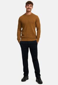 Mustard knit sweater with herringbone pattern, ribbed cuffs and hem, paired with dark pants and black shoes. Model stands with hands in pockets.