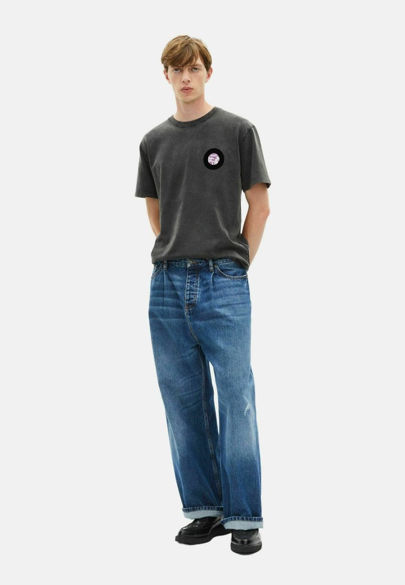 Grå bomulds t-shirt med et cirkulært grafisk motiv på brystet, paired med løse blå denim jeans og sorte sko.