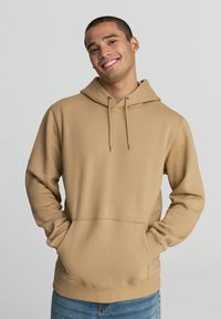 Felpa cappuccio color cammello con tasca frontale, cappuccio con lacci, polsini e orlo a coste. Texture liscia e vestibilità rilassata, abbinata a jeans blu.