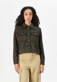 Vrouw met lang krullend haar, gekleed in een donkergroene leopardprint blouse met knopen en beige cargobroek, staande tegen een witte achtergrond.