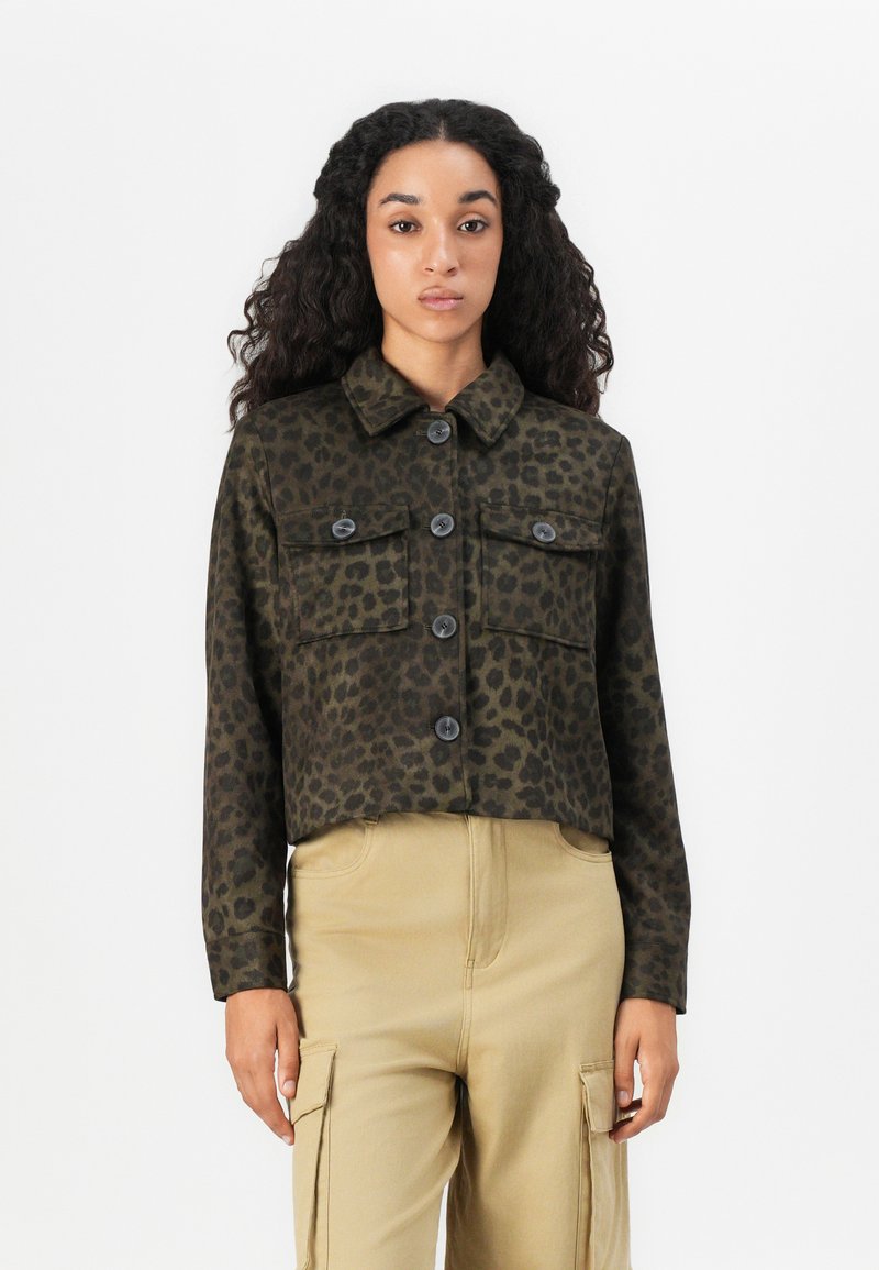 Vrouw met lang krullend haar, gekleed in een donkergroene leopardprint blouse met knopen en beige cargobroek, staande tegen een witte achtergrond.