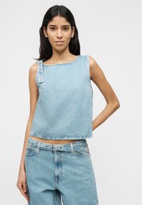 Jeune femme portant un haut sans manches en denim bleu clair avec détail de boucle et un jean taille haute assorti, debout devant un fond blanc uni.