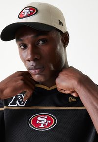 Black mesh jersey with gold accents, featuring a red and white SF logo. Beige cap with a red and white embroidered logo. Model adjusting collar.