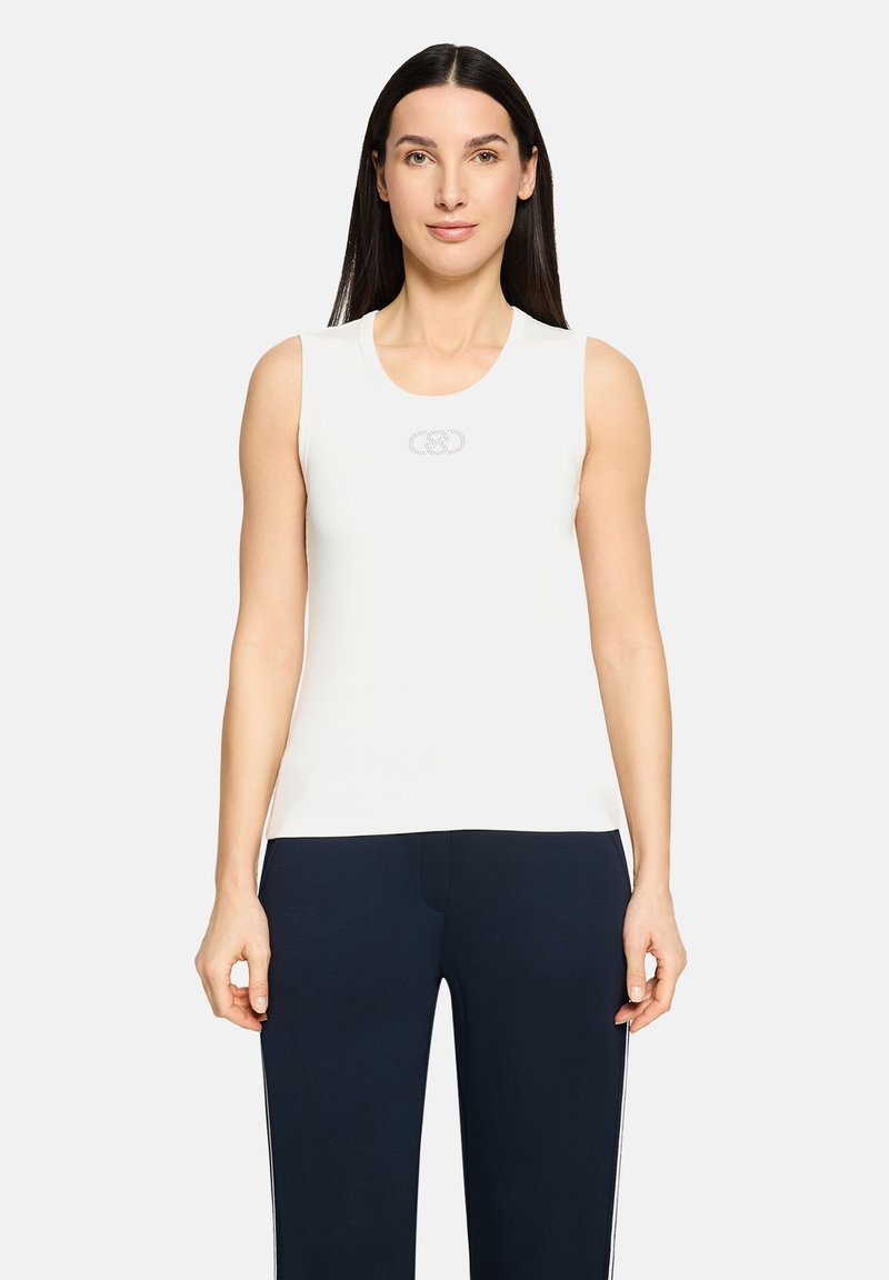 Haut blanc sans manches avec un col rond, arborant un logo discret au centre. Tissu lisse et coupe ajustée, associé à un pantalon foncé.