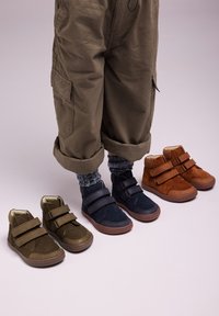 Un par de botas de ante en verde oliva, azul marino y marrón con correas de velcro. Las botas tienen punteras redondas y suelas de goma, mostradas junto a pantalones doblados.