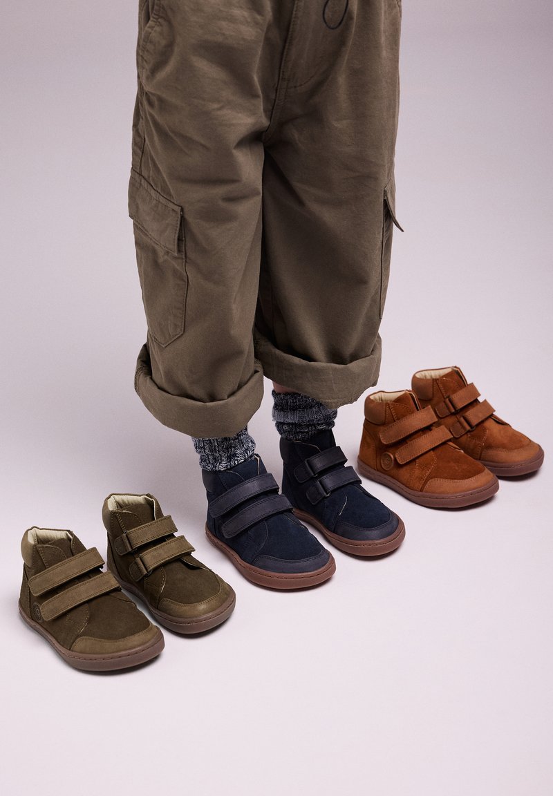 Un par de botas de ante en verde oliva, azul marino y marrón con correas de velcro. Las botas tienen punteras redondas y suelas de goma, mostradas junto a pantalones doblados.