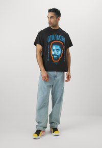 Sort bomulds T-shirt med et blåt og orange grafik af Clyde Frazier's ansigt, samt fed tekst. Matcher med løse lyseblå jeans og multifarvede sneakers.