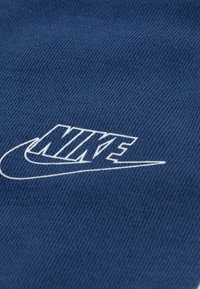 Μπλε βαμβακερό ύφασμα με υφασμένη υφή που παρουσιάζει το λευκό λογότυπο της Nike σε στιλιζαρισμένη γραμματοσειρά. Ο σχεδιασμός είναι κοντινός και λεπτομερής.