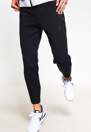 Person, die schwarze Nike-Jogginghosen und weiße Nike-Sneaker trägt, macht einen Schritt nach vorne vor einem schlichten weißen Hintergrund.