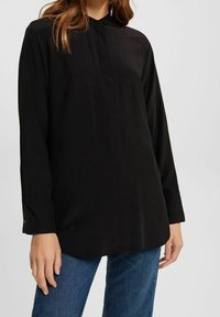 Maglietta nera a maniche lunghe con colletto bottonato, texture liscia e vestibilità ampia, abbinata a jeans in denim blu.