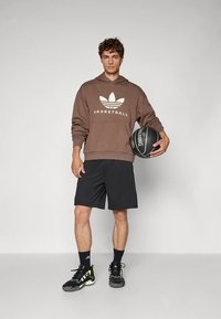 Brown hoodie with white "BASKETBALL" text and logo, black shorts, black sneakers with green accents, holding a black basketball.