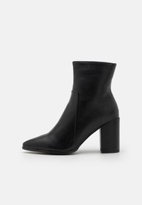 Botte cheville en cuir noir avec bout pointu et talon bloc épais sur un fond gris clair uni.