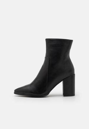 Botte cheville en cuir noir avec bout pointu et talon bloc épais sur un fond gris clair uni.