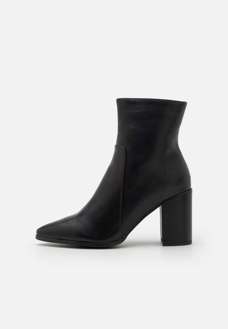 Botte cheville en cuir noir avec bout pointu et talon bloc épais sur un fond gris clair uni.