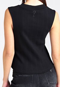 Mouwloos zwart geribbeld topje met een hoge halslijn, voorzien van een klein logo aan de achterkant en een nauwsluitend ontwerp, gecombineerd met een zwarte spijkerbroek.