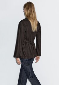 Braune Jacke mit Gürtel, langen Ärmeln, lockerer Passform und strukturiertem Stoff, getragen mit dunklen Jeans. Accessoires und Muster nicht sichtbar.