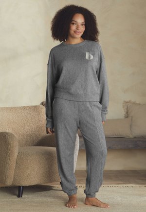 Žena s kučeravými vlasmi, ktorá má na sebe sivý pletený top s dlhým rukávom a zodpovedajúce nohavice, stojí bosá v útulnej obývacej izbe s béžovým kreslom a vankúšmi.