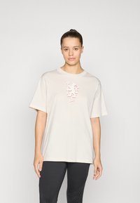 Beige t-paita, jossa on lyhyet hihat ja kohotettu punainen graafinen muotoilu rinnassa. Yhdistetty tummiin legginsseihin.