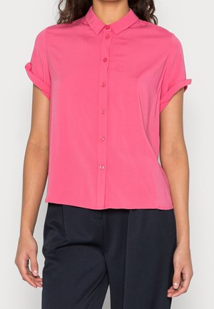 Nainen, joka pukeutunut lyhythihaiseen vaaleanpunaiseen napitettavaan paitaan, jonka hihat on kääritty, ja mustiin housuihin, näkyy kaulasta lantioihin asti.