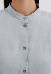Blouse en lin gris clair avec un col montant, dotée d'une patte de boutonnage et d'un tissu texturé. Les boutons sont ronds et gris foncé.