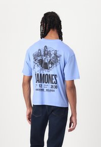 Light blue cotton t-shirt with short sleeves. Graphic print on the back featuring band members, text, and event details in black.