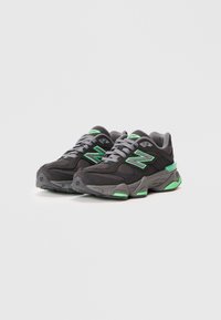 Mustunud musta ja halli värvi New Balance'in ketsid roheliste aktsentide ja nööpidega, näidatud kõrvuti lihtsal valgel taustal.
