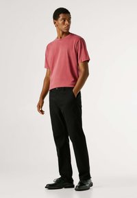 Rood katoenen t-shirt gecombineerd met zwarte broek. Model staat met handen in de zakken, draagt zwarte schoenen. Eenvoudig ontwerp, geen zichtbare patronen.