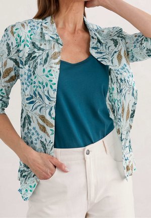 Kobieta nosząca turkusowy top na ramiączkach z okrągłym dekoltem, jasnoniebieską, kwiecistą koszulę z podwiniętymi rękawami oraz kremowe spodnie z wysokim stanem.