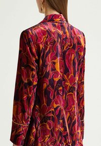 Blouse à manches longues en tissu velours avec un motif floral aux nuances de rose, violet et orange. Comprend un col ajusté et des détails de boutons.